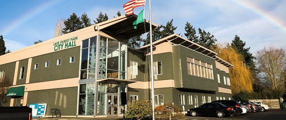 Lake Forest Park city hall with a rainbow overhead; US flag flying, several cars parked in the parking lot